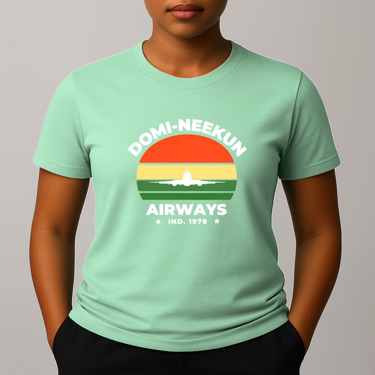 Person wearing a mint green short-sleeve T-shirt with a retro-style graphic reading 'DOMI-NEEKUN AIRWAYS' and 'IND. 1978' over a striped sunset and airplane silhouette.