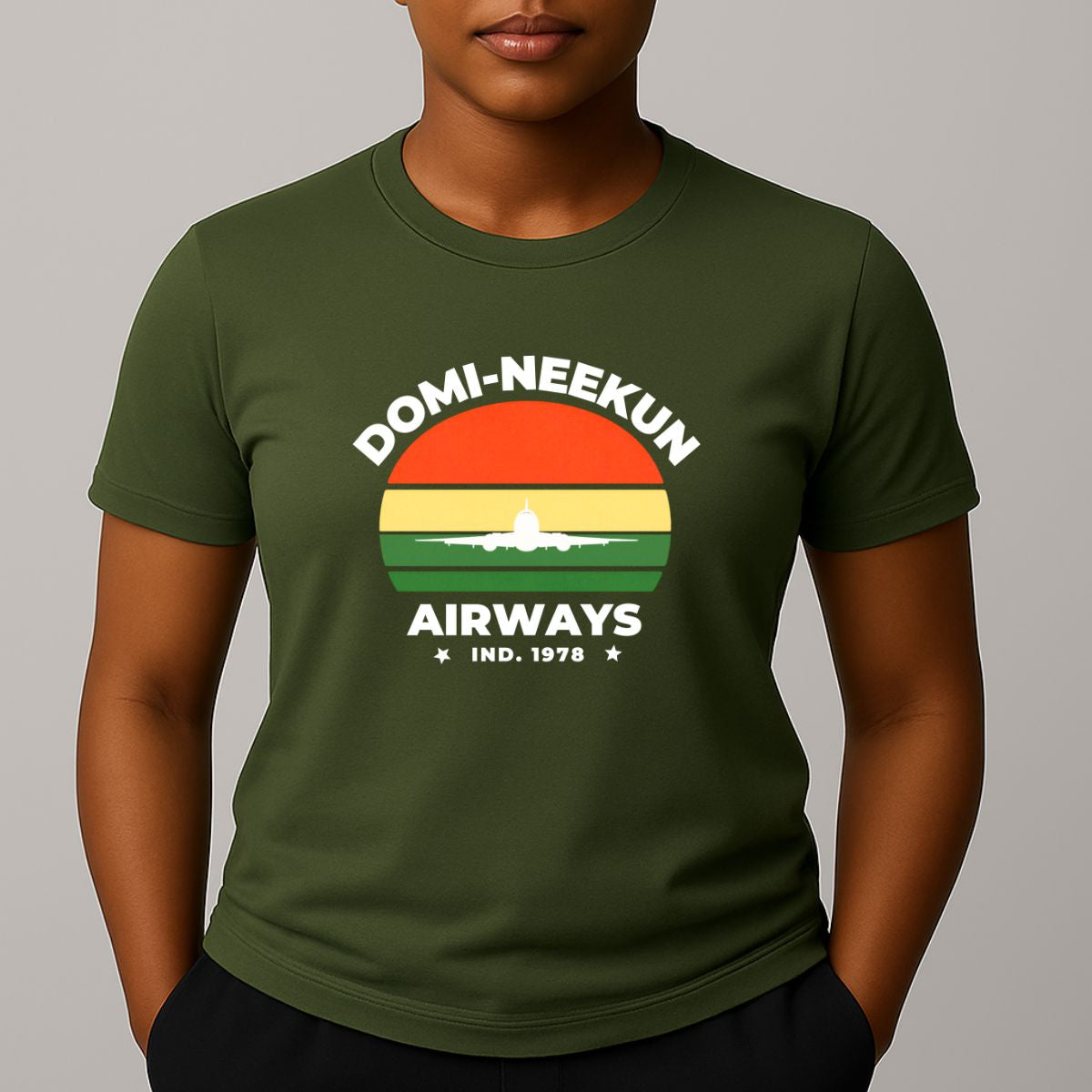 Person wearing a khaki green short-sleeve T-shirt with a retro-style graphic reading 'DOMI-NEEKUN AIRWAYS' and 'IND. 1978' over a striped sunset and airplane silhouette.