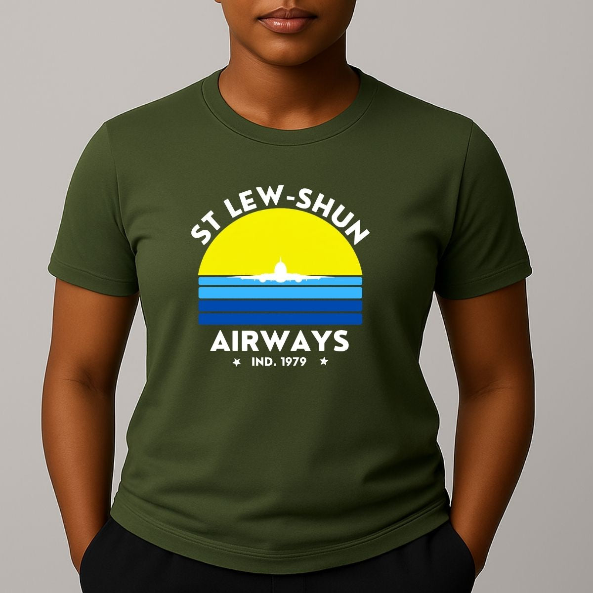 Person wearing an khaki green t-shirt with a graphic print that reads 'ST LEW-SHUN AIRWAYS IND. 1979' in white, above and below a colourful striped circle with a white airplane in the center.